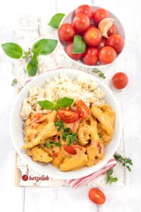 Putenpfanne mit Reis nach einem Putengeschnetzeltes mit Tomaten-Frischkäse-Soße Rezept von herzelieb auf einem weißen Teller mit einer Portion Reis. Die zarten Putenstreifen in der samtigen Soße sind mit Basilikum und Thymian dekoriert. Frische Tomaten und Knoblauch als Zutaten im Hintergrund.
