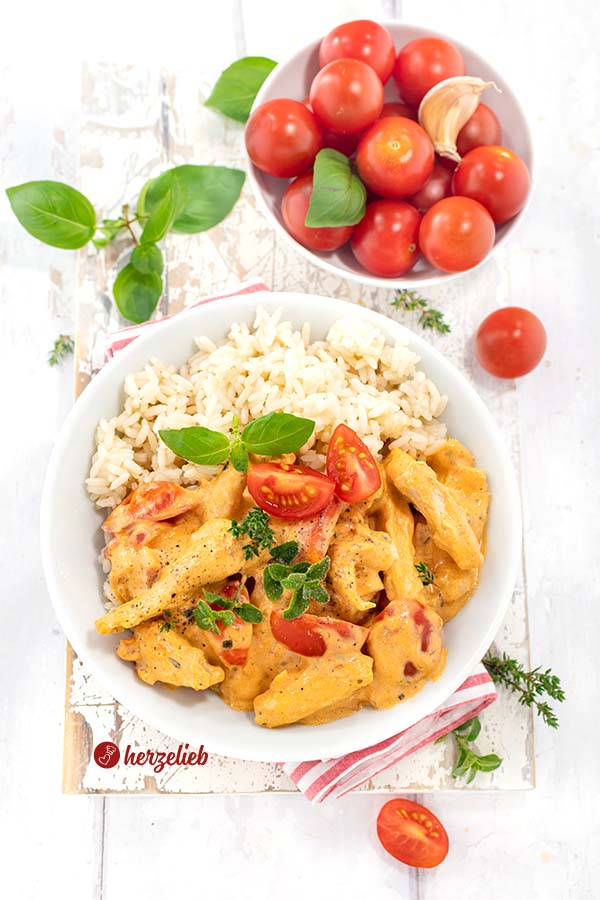 Putenpfanne mit Reis nach einem  Putengeschnetzeltes mit Tomaten-Frischkäse-Soße Rezept von herzelieb auf einem weißen Teller mit einer Portion Reis. Die zarten Putenstreifen in der samtigen Soße sind mit Basilikum und Thymian dekoriert.  Frische Tomaten und Knoblauch als Zutaten im Hintergrund.