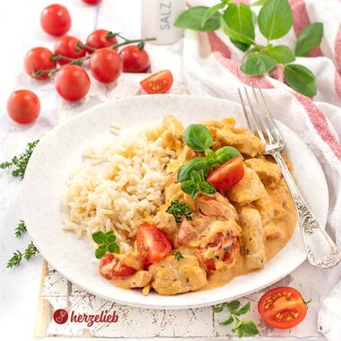 Schnelle Putenpfanne nach einem Putengeschnetzeltes mit Tomaten-Frischkäse-Soße Rezept von herzelieb, angerichtet auf einem weißen Teller, der neben Reis auch frische Tomatenstücke und Kräuter enthält. Rundherum liegen weitere Zutaten wie Kirschtomaten, Basilikum und eine Salzstreuer.