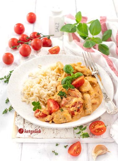 Schnelle Putenpfanne nach einem Putengeschnetzeltes mit Tomaten-Frischkäse-Soße Rezept von herzelieb, angerichtet auf einem weißen Teller, der neben Reis auch frische Tomatenstücke und Kräuter enthält. Rundherum liegen weitere Zutaten wie Kirschtomaten, Basilikum und eine Salzstreuer.