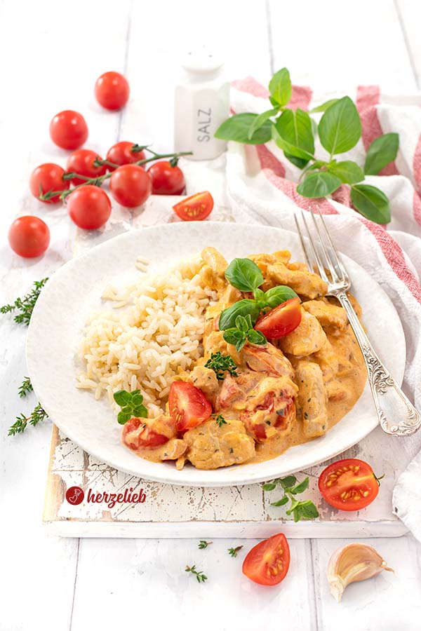 Schnelle Putenpfanne nach einem Putengeschnetzeltes mit Tomaten-Frischkäse-Soße Rezept von herzelieb, angerichtet auf einem weißen Teller, auf dem neben Reis auch frische Tomatenstücke und Kräuter sind. Rundherum liegen weitere Zutaten wie Kirschtomaten, Basilikum und ein Salzstreuer.