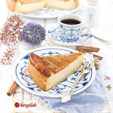 Ein einzelnes Stück Saure-Sahne-Kuchen nach einem Rahmkuchen Rezept von herzelieb, serviert auf einem blau-weiß gemusterten Teller mit einer Gabel daneben und einer Tasse Kaffee im Hintergrund. Dekoriert mit Blumen und Zimtstangen. Im Hintergrund der angeschnittene Kuchen auf eine Platte.