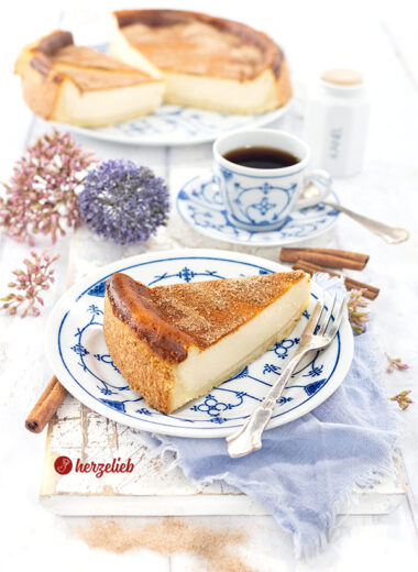 Ein einzelnes Stück Saure-Sahne-Kuchen nach einem Rahmkuchen Rezept von herzelieb, serviert auf einem blau-weiß gemusterten Teller mit einer Gabel daneben und einer Tasse Kaffee im Hintergrund. Dekoriert mit Blumen und Zimtstangen. Im Hintergrund der angeschnittene Kuchen auf eine Platte.