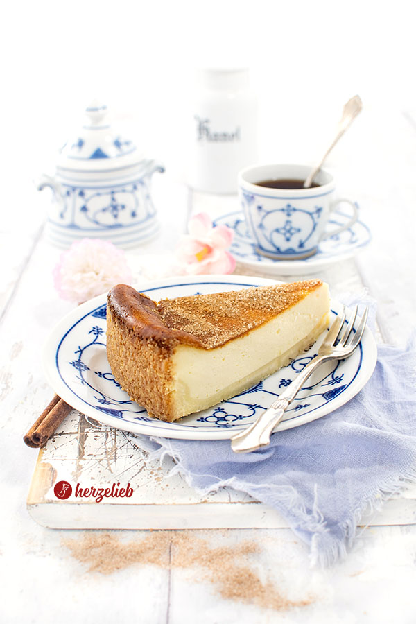 Ein Stück Saure-Sahne-Kuchen mit Zimtzucker nach einem Rahmkuchen Rezept von herzelieb auf einem Teller mit einer Kuchengabel, daneben und dahinter eine Tasse Kaffee und ein Zuckerdöschen. Dekoriert mit Zimtstangen und Blumen.
