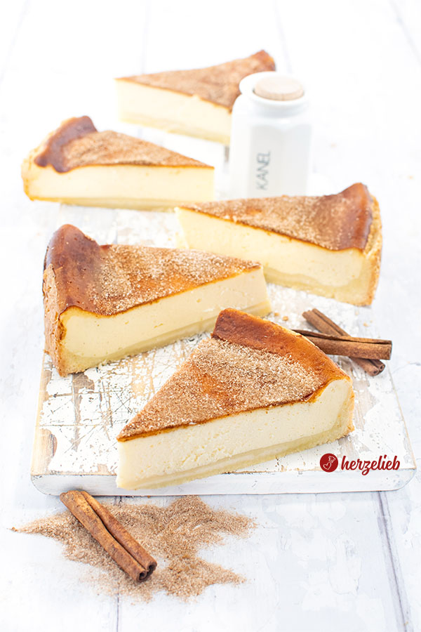 Mehrere Stücke Saure-Sahne-Kuchen nach einem Rahmkuchen Rezept von herzelieb auf einer weißen Holzplatte angerichtet, umgeben von Zimtstangen und Zimtpulver, im Hintergrund ein Gewürzbehälter