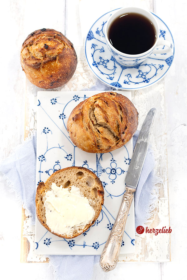 Zwei goldbraune Röstzwiebelbrötchen mit Roggenmehl auf einem weiß-blauem Porzellanbrett. Ein Brötchen ist aufgeschnitten und mit Butter bestrichen, daneben ein Messer und oben rechts eine Tasse Kaffee.