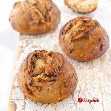 Drei frisch gebackene Röstzwiebelbrötchen mit knuspriger, goldbrauner Kruste liegen auf einem weißen, leicht abgenutzten Holzbrett.