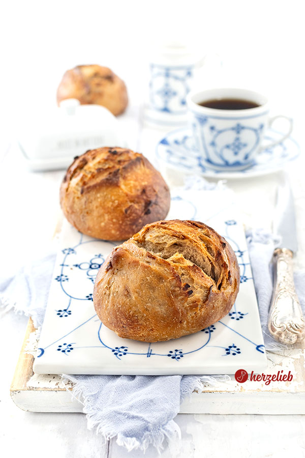 Zwei frisch gebackene Röstzwiebelbrötchen mit Roggenmehl liegen auf einem blau-weiß dekorierten Porzellanbrett, daneben ein Silbermesser. Im Hintergrund eine Tasse Kaffee, eine Butterglocke, ein Milchkännchen und ein weiteres Brötchen.