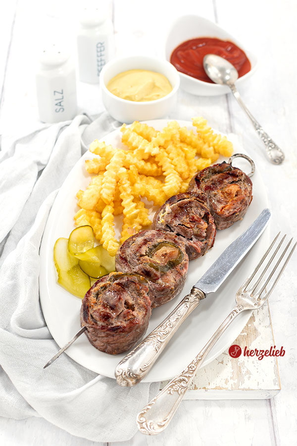 Ein Grillspieß mit 4 Rouladenscheiben nach einem Rouladenspieße Rezept von herzelieb. Angerichet mit Pommes. Vorne auf dem Tellerrand liegt Silberbesteck. Im Hintergrund Senf, Ketchup und ein Gewürzstreuer.