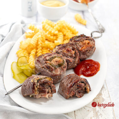 Grillspieße nach einem Rouladenspieße Rezept von herzelieb. Angerichtet auf einem weißen Teller mit Pommes und Ketschup. Im Hintergund ein Gewürzstreuer, ein Schälchen mit Senf und eines mit Ketchup.