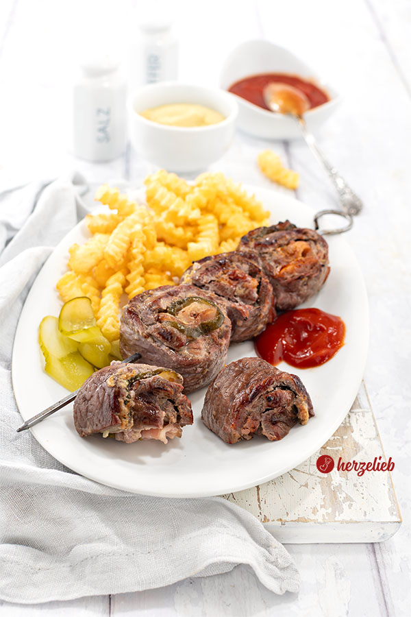 Grillspieße nach einem Rouladenspieße Rezept von herzelieb. Angerichtet auf einem weißen Teller mit Pommes und Ketschup. Im Hintergund ein Gewürzstreuer, ein Schälchen mit Senf und eines mit Ketchup.