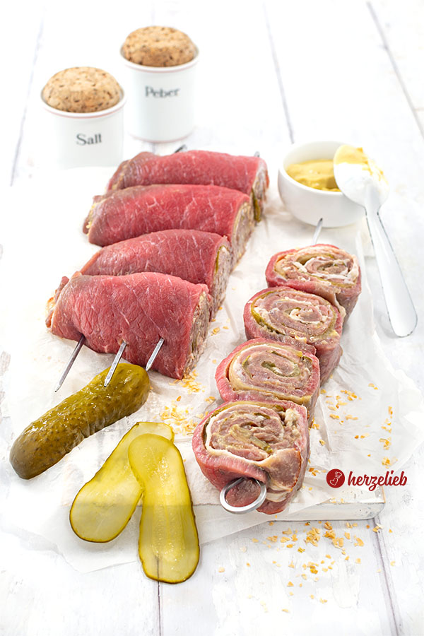 Rouladenspieße vom Grill in der Vorbereitung. Links die aufgerollten Fleischstücke mit den Spießen, rechts ein abgeschnittener Spieß. Dahinter eine Schale Senf. Weiter hinten 2 Gewürzbehälter, vorne links eine längs halbierte Gurke und 2 Gurkenscheiben.