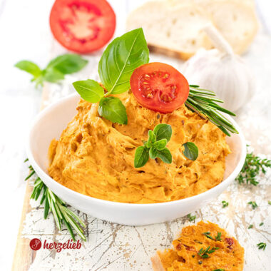 Eine weiße Schale gefüllt mit cremiger Tomatenbutter mit Parmesan, garniert mit frischen Basilikumblättern, einem Tomatenscheibe und Rosmarinzweigen, im Hintergrund liegen Knoblauch und Brot auf einem weißen Holzbrett.