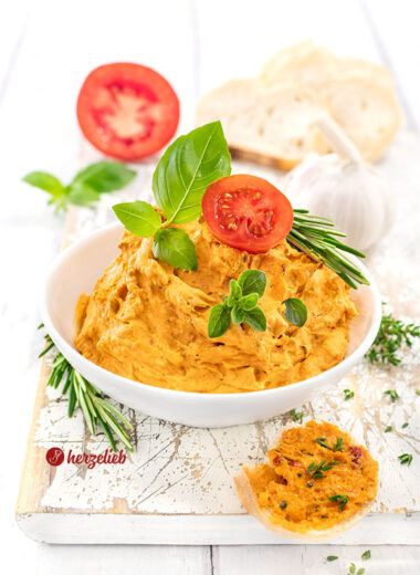 Eine weiße Schale gefüllt mit cremiger Tomatenbutter mit Parmesan, garniert mit frischen Basilikumblättern, einem Tomatenscheibe und Rosmarinzweigen, im Hintergrund liegen Knoblauch und Brot auf einem weißen Holzbrett.