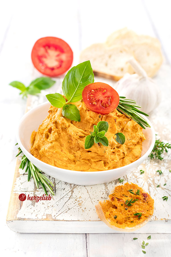 Eine weiße Schale gefüllt mit cremiger Tomatenbutter mit Parmesan, garniert mit frischen Basilikumblättern, einem Tomatenscheibe und Rosmarinzweigen, im Hintergrund liegen Knoblauch und Brot auf einem weißen Holzbrett.