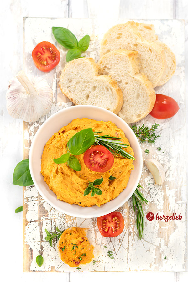 Von oben aufgenommenes Bild einer Schale mit würziger Tomatenbutter, dekoriert mit frischen Kräutern und einer Tomatenscheibe, umgeben von Knoblauchzehen und mehreren Scheiben Weißbrot auf einer weißen, rustikalen Holzunterlage.