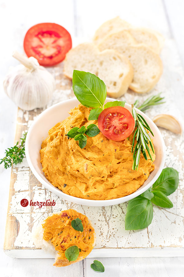 Rote Kräuterbutter mit getrockneten Tomaten, und Parmesan nach einem Tomatenbutter Rezept von herzelieb. Angerichtet in einer weißen Schale. Dekoriert mit Tomate, Oregano, Basilikum und Rosmarin. Im Hintergrund Brot, Knoblauch und Tomaten.