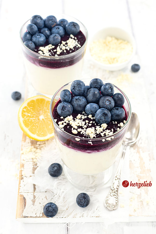 Zwei Dessertgläser mit Blaubeer-Käsekuchencreme im Glas. Unten im Glas die helle Creme, darauf Blaubeersoße und darauf frische Blaubeeren und weiße Schokoladenrapsel. Neben dem vorderen Glas eine halbe Zitrone. Hinten rechts ein Schälchen mit gerapselter Schokolade.