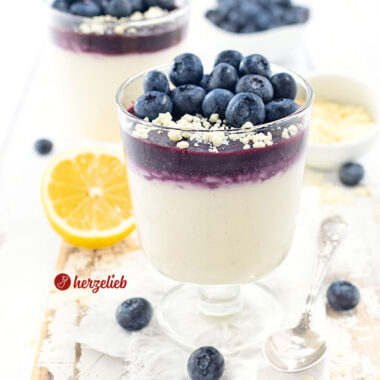 2 Gläser mit Blaubeer-Käsekuchencreme Dessert im Glas. Unten im Glas ist die helle Creme, darüber das Blaubeermus und darauf Schokoraspeln und frische Blaubeeren. Im hinergrund eien Zitrone,, eine Schale mit weißen Schokoraspeln und ein Schälchen mit Blaubeeren.