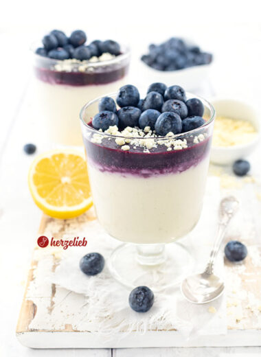 2 Gläser mit Blaubeer-Käsekuchencreme Dessert im Glas. Unten im Glas ist die helle Creme, darüber das Blaubeermus und darauf Schokoraspeln und frische Blaubeeren. Im hinergrund eien Zitrone,, eine Schale mit weißen Schokoraspeln und ein Schälchen mit Blaubeeren.