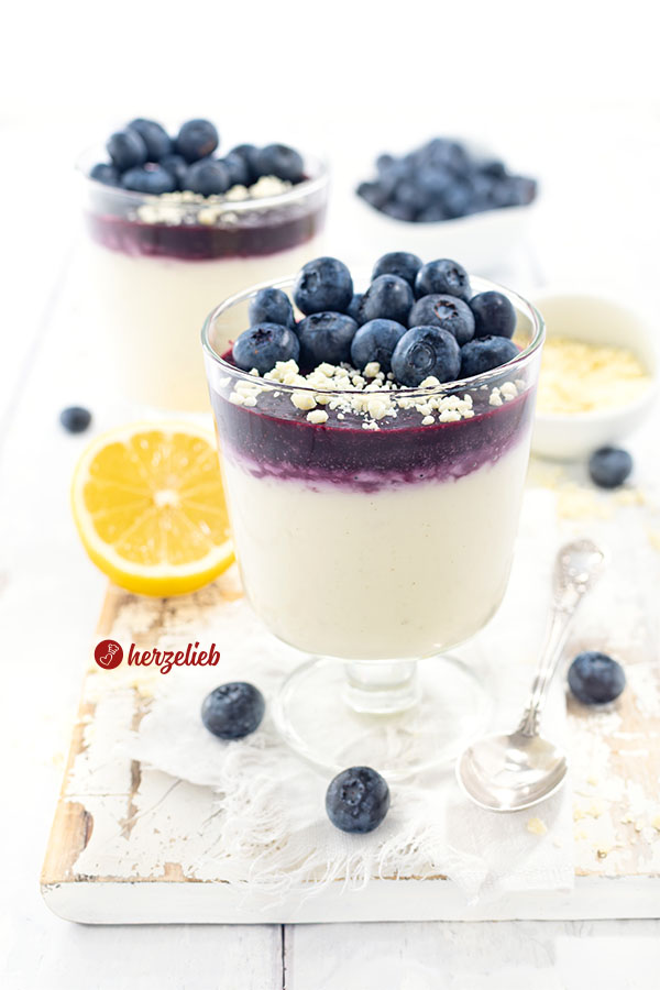 2 Gläser mit Blaubeer-Käsekuchencreme Dessert im Glas. Unten im Glas ist die helle Creme, darüber das Blaubeermus und darauf Schokoraspeln und frische Blaubeeren. Im Hintergrudn eien Zitrone, eine Schale mit weißen Schokoraspeln und ein Schälchen mit Blaubeeren. 