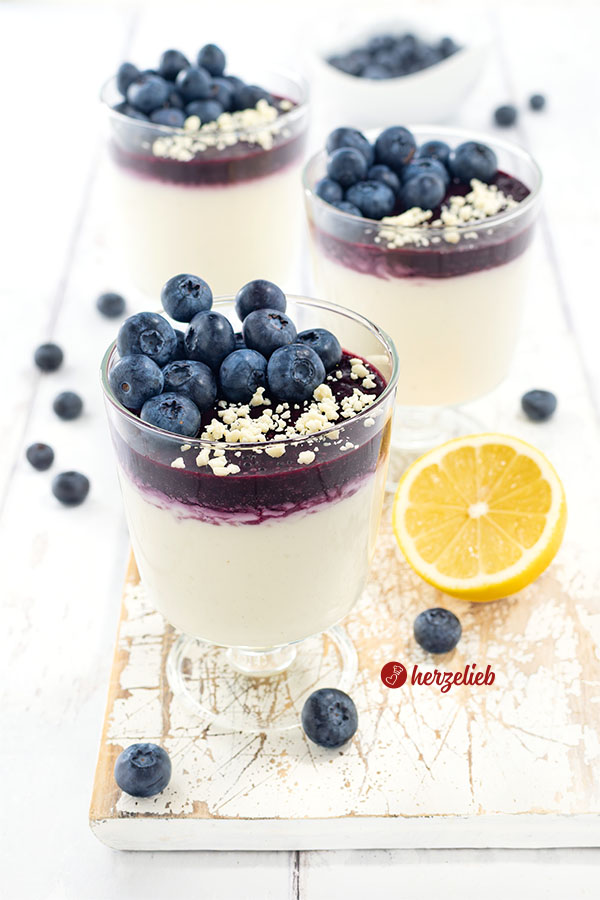 Drei Gläser mit einem Nachtisch nach einem Blaubeer-Käsekuchencreme Rezept von herzelieb auf einem weißen Holzbett.Im Glas unten die helle Creme, darüber eine Schicht Blaubeermus. Obenauf dekoriert mit frischen Blaubeeren und geraspelter weißer Schokolade. 
