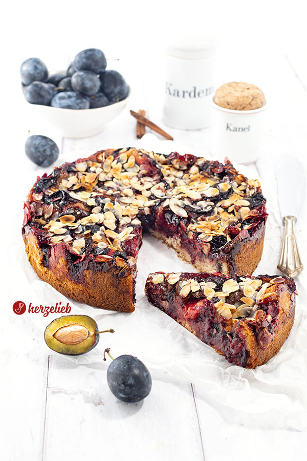 Ein angeschnittener Zwetschgen-Butterkuchen mit Mandeln nach einem Blommekage Rezept aus Dänemark von herzelieb Der Pflaumenkuchen liegt auf Backpapier, ein Stück ist nach vorne gezogen. Vor und hinter dem Kuchen liegen Zwetschgen. Rechts neben dem Kuchen ein Kuchenmesser