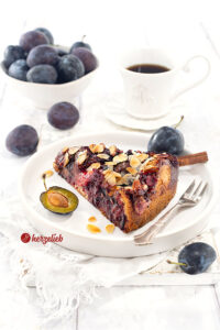 Ein Stück Blommekage, der Zwetschgen-Butterkuchen mit Hefeteig und Butterguss aus Dänemark auf einem kleinen Teller. Auf dem Tellerrand eine Kuchengabel.Hinter dem Pflaumenkuchen mit Mandeln eine Tasse Kaffee und eine Schale mit Pflaumen.