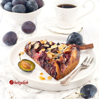 Ein Stück Blommekage, der Zwetschgen-Butterkuchen mit Hefeteig und Butterguss aus Dänemark auf einem kleinen Teller. Auf dem Tellerrand eine Kuchengabel.Hinter dem Pflaumenkuchen mit Mandeln eine Tasse Kaffee und eine Schale mit Pflaumen.