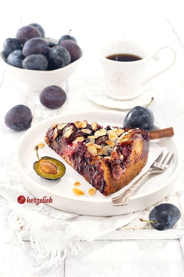 Ein Stück Blommekage, der Zwetschgen-Butterkuchen mit Hefeteig und Butterguss aus Dänemark auf einem kleinen Teller. Auf dem Tellerrand eine Kuchengabel.Hinter dem Pflaumenkuchen mit Mandeln eine Tasse Kaffee und eine Schale mit Pflaumen.