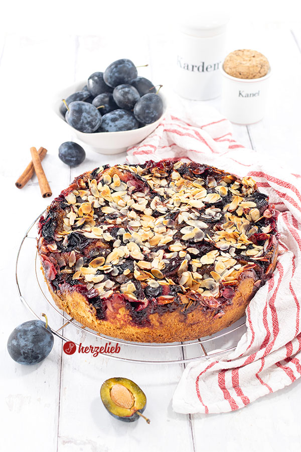 Ein Stück Blommekage, der Zwetschgen-Butterkuchen mit Hefeteig und Butterguss aus Dänemark auf einem kleinen Teller. Auf dem Tellerrand eine Kuchengabel.Hinter dem Pflaumenkuchen mit Mandeln eine Tasse Kaffee und eine Schale mit Pflaumen.   Ein ganzer Blommekage auf einem Backgitter. Der Zwetschgen-Butterkuchen aus Dänemark ist mit Mandelblättchen bestreut. Rechts vom Kuchen liegt ein weißes Geschirrtuch mit roten Streifen. Hinter dem Pflaumenkuchen steht eine Schale mit weiteren Zwetschgen sowie zwei Gewürzgefäße