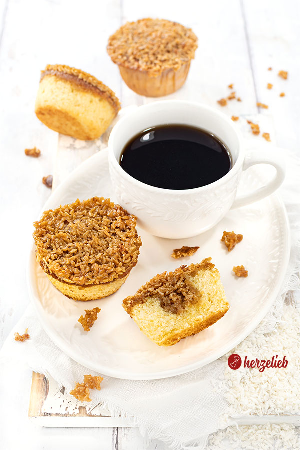 Ein weißer, ovaler Teller mit kleinen einem ganzen und einem halben Kuchen nach einem Drømmekage Muffins Rezept aus Dänemark von herzelieb. Bei dem Muffin vorne recht sieht man die Füllung. Im Hintergrund zwei weitere Muffins. Alles ist angerichtet auf einem weißen Holzbrett.