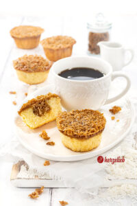 Mehrere kleine Kuchen nach einem Drømmekage Muffins Rezept aus Dänemark von herzelieb, davon einer aufgeschnitten, der die karamellige Kokosfüllung sichtbar macht, dazu eine weiße Kaffeetasse und verstreute Haferflocken einem weißen, ovalen Teller. Der Teller steht auf eiinem weißen Holzbrett, das auf einem weißen Leinentuch liegt.