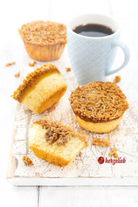 Drei ganze Drømmekage Muffins mit knuspriger Karamellkruste auf einem weißen Holzbrett mit einem blauen Becher Kaffee. Im Vordergrund liegt ein halber Muffin.