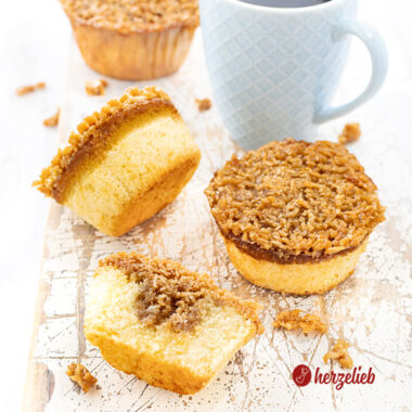 Drei ganze Drømmekage Muffins mit knuspriger Karamellkruste auf einem weißen Holzbrett mit einem blauen Becher Kaffee. Im Vordergrund liegt ein halber Muffin.