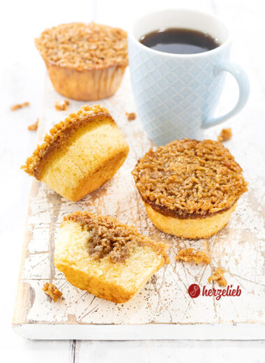 Drei ganze Drømmekage Muffins mit knuspriger Karamellkruste auf einem weißen Holzbrett mit einem blauen Becher Kaffee. Im Vordergrund liegt ein halber Muffin.