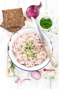 Cremiger Skinkesalat nach einem Fuglekvidder Rezept von herzelieb in einer weißen Emailleschüssel mit blauem Rand. Garniert mit fein geschnittenem Schnittlauch und roten Zwiebelringen, daneben zwei Scheiben dunkles Vollkornbrot und eine rote Zwiebel auf einem hellen Holzbrett.