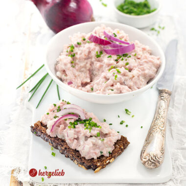 Das Bild zeigt eine Portion dänischen Skinkesalat nach einem Fuglekvidder Rezept von herzelieb auf einem knusprigen Roggenbrot, dekoriert mit roten Zwiebelringen und fein gehacktem Schnittlauch,. Im Hintergrund steht eine Schale mit dem Brotaufstrich. Dahinter liegt eine ganze rote Zwiebel und ein kleines Schälchen mit Schnittlauchröllchen.