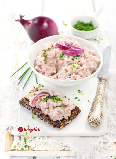 Das Bild zeigt eine Portion dänischen Skinkesalat nach einem Fuglekvidder Rezept von herzelieb auf einem knusprigen Roggenbrot, dekoriert mit roten Zwiebelringen und fein gehacktem Schnittlauch,. Im Hintergrund steht eine Schale mit dem Brotaufstrich. Dahinter liegt eine ganze rote Zwiebel und ein kleines Schälchen mit Schnittlauchröllchen.