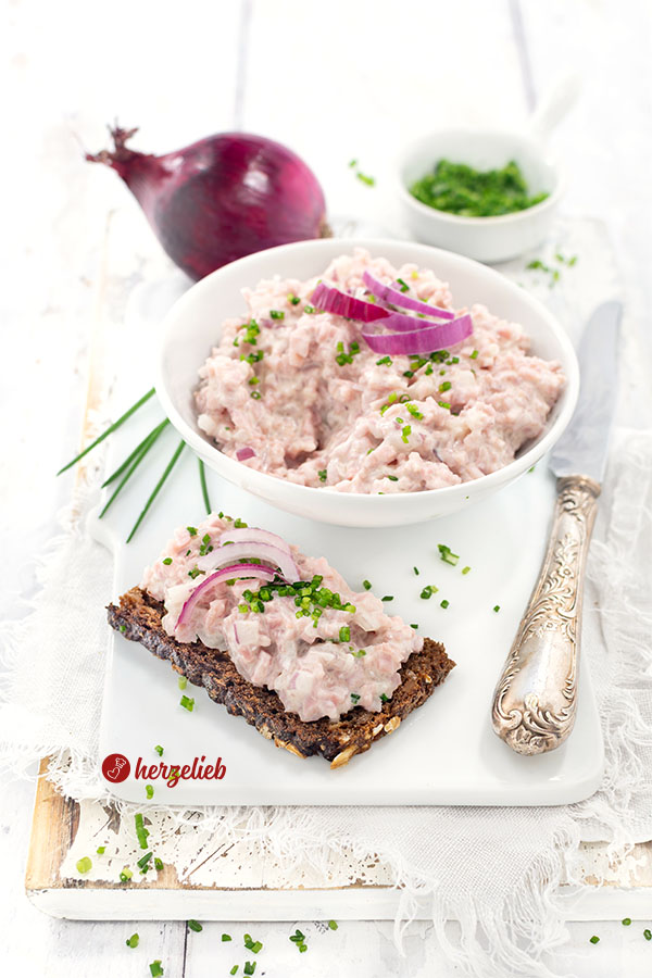 Das Bild zeigt eine Portion dänischen Skinkesalat nach einem Fuglekvidder Rezept von herzelieb auf einem knusprigen Roggenbrot, dekoriert mit roten Zwiebelringen und fein gehacktem Schnittlauch,. Im Hintergrund steht eine Schale mit dem Brotaufstrich. Dahinter liegt eine ganze rote Zwiebel und ein kleines Schälchen mit Schnittlauchröllchen.