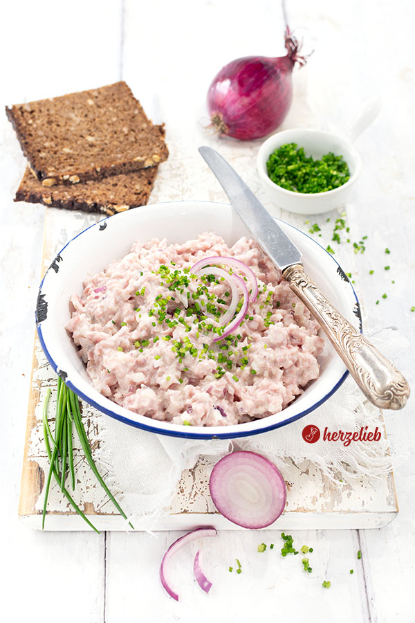 Eine Schüssel mit cremigem Fuglekvidder, einem dänischen Skinkesalat, garniert mit frisch geschnittenem Schnittlauch und roten Zwiebelringen, steht auf einem weißen Holzbrett vor zwei Scheiben Vollkornbrot, einer roten Zwiebel und einem Schälchen Schnittlauchröllchen.