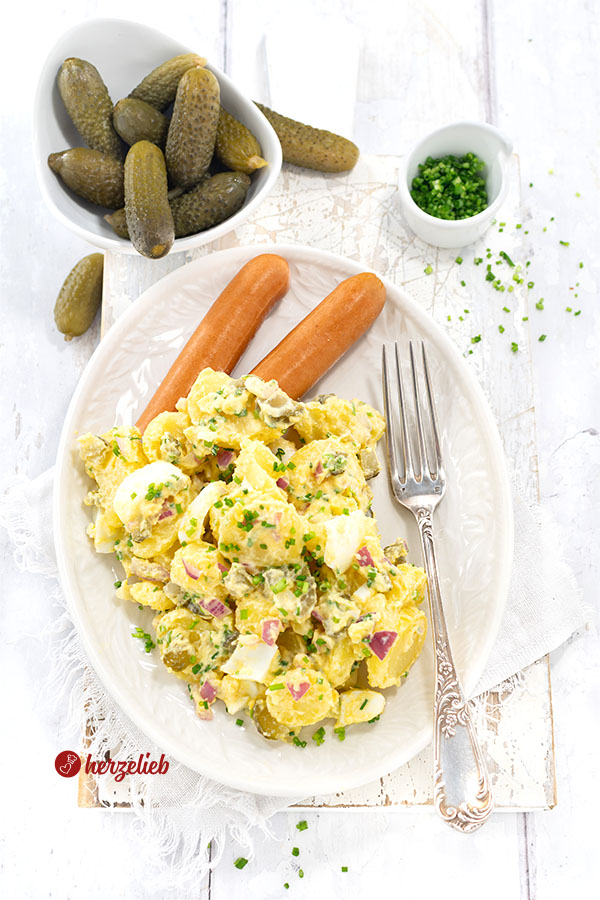 Ein Teller mit Salat nach einem gekochter Kartoffelsalat Rezept von von herzelieb. Serviert mit Würstchen, dekoriert mit Schnittlauchröllchen, auf dem Tellerrand liegt eine Gabel. Im Hintergrund eine rote Zwiebel, eine Schale Gewürzgurken udn ein Schälchen mit Schnittlauch