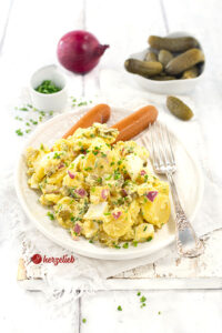 Gekochter Kartoffelsalat auf einem weißen, ovalen Teller mit einer Silbergabel auf dem Tellerrand. Serviert mit Würstchen, dekoriert mit Schnittlauchröllchen. Im Hintergrund Gewürzgurken, eine rote Zwiebel und ein Schälchen mit gehacktem Schnittlauch