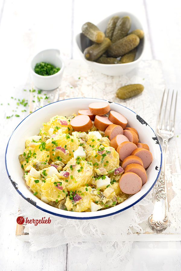 In einer weißen Emailleschale mit blauem Rand ist gekochter Kartoffelsalat mit falscher Mayonnaise angerichtet. Am Schüsselrand liegen Würstchenscheiben, der Salat ist mit Schnittlauchröllchen bestreut. Rechts neben der Schüssel liegt eine Gabel. Im Hintergrund Cornichons und eine kleine Schale mit Schnittlauch