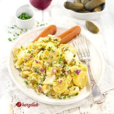 Gekochter Kartoffelsalat auf einem weißen, ovalen Teller mit einer Silbergabel auf dem Tellerrand. Serviert mit Würstchen, dekoriert mit Schnittlauchröllchen. Im Hintergrund Gewürzgurken, eine rote Zwiebel und ein Schälchen mit gehacktem Schnittlauch