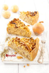 3 Stücke goldgelb gebackener Kohl-Kartoffel-Kuchen auf einem weißen Holzbrett. Rechts vom vorderen Stück eine Gabel. Im Hintergrund eine Zwiebel und drei Kartoffeln.