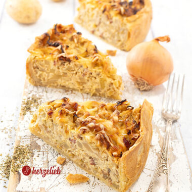 3 Stücke goldgelb gebackener Kohl-Kartoffel-Kuchen auf einem weißen Holzbrett. Rechts vom vorderen Stück eine Gabel. Im Hintergrund eine Zwiebel und drei Kartoffeln.
