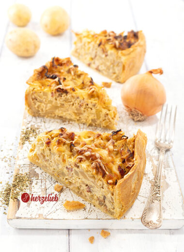 3 Stücke goldgelb gebackener Kohl-Kartoffel-Kuchen auf einem weißen Holzbrett. Rechts vom vorderen Stück eine Gabel. Im Hintergrund eine Zwiebel und drei Kartoffeln.