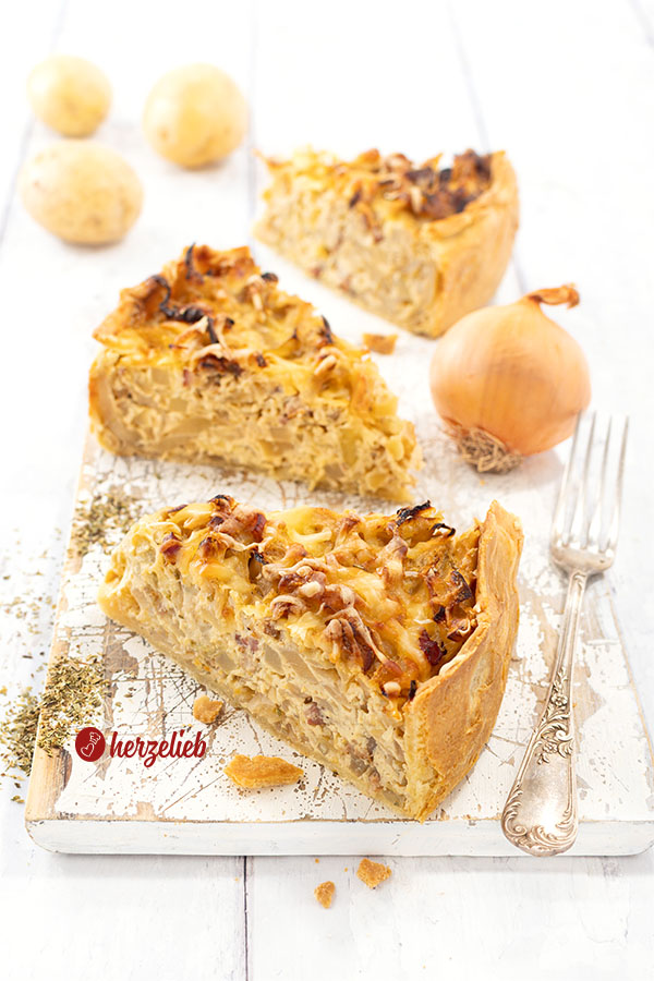 3 Stücke goldgelb gebackener Kohl-Kartoffel-Kuchen auf einem weißen Holzbrett. Rechts vom vorderen Stück eine Gabel. Im Hintergrund eine Zwiebel und drei Kartoffeln. 