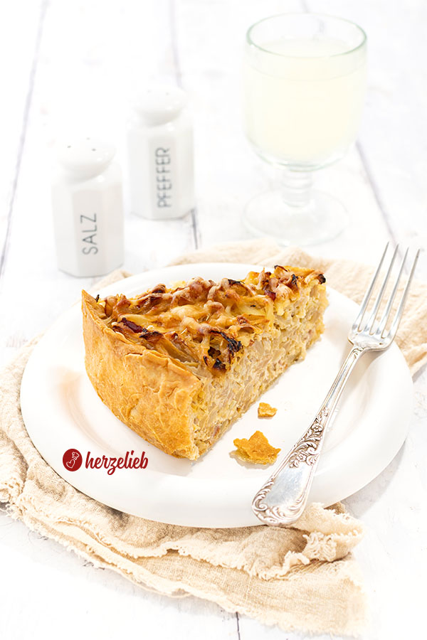 Ein Stück herzhafter, goldraun gebackener Kuchen nach einem Kohl-Kartoffel-Kuchen Rezept von herzelieb auf einem weißen Teller, auf dessen Rand eine Gabel liebt. im Hintergrund ein Salz- und ein Pfefferstreuser und ein Glas Wein.